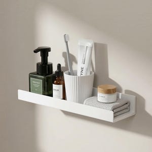 Op de afbeelding: Een witte badkamerplank met diverse toiletartikelen. Een donkergroene zeepdispenser, een bruine serumfles, een tandenborstel, tandpasta, een klein potje balsem en een gevouwen grijze washand zijn op de plank geplaatst.
