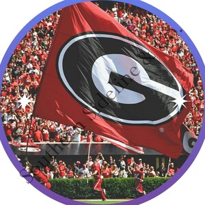 K&ouml;nnte beinhalten: Eine rote Flagge mit einem schwarz-wei&szlig;en "G"-Logo wird von einer Person in roter Uniform gehalten, mit einer Menschenmenge im Hintergrund. Die Flagge weht im Wind, und das Logo ist deutlich sichtbar.