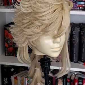 May include: A light blonde wig styled with layered bangs and a long, braided ponytail. The wig is displayed on a mannequin head attached to a black stand. Bookshelves filled with books and decorative items are in the background.