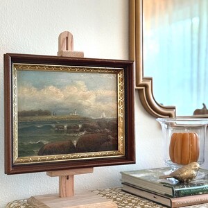 May include: A landscape painting in a dark brown and gold frame, displayed on a light-colored wooden easel. The artwork depicts a coastal scene with a lighthouse on a rocky shore, a sailboat on the water, and a cloudy blue sky above the horizon.