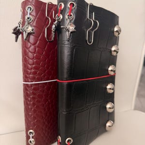 May include: Two leather journal covers, one burgundy and one black, are displayed side-by-side. The burgundy cover has a textured, scale-like pattern, while the black cover has a grid pattern. Both have metal accents and decorative charms.