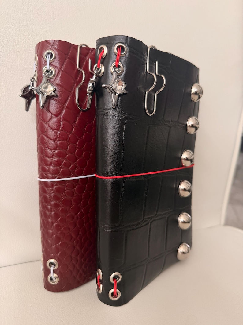 May include: Two leather journal covers, one burgundy and one black, are displayed side-by-side. The burgundy cover has a textured, scale-like pattern, while the black cover has a grid pattern. Both have metal accents and decorative charms.