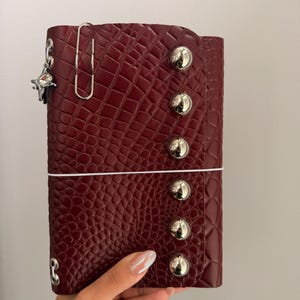 May include: A burgundy faux-leather planner with a crocodile skin texture. The planner has silver metal button accents and a silver paper clip. A small star-shaped charm hangs from the planner.