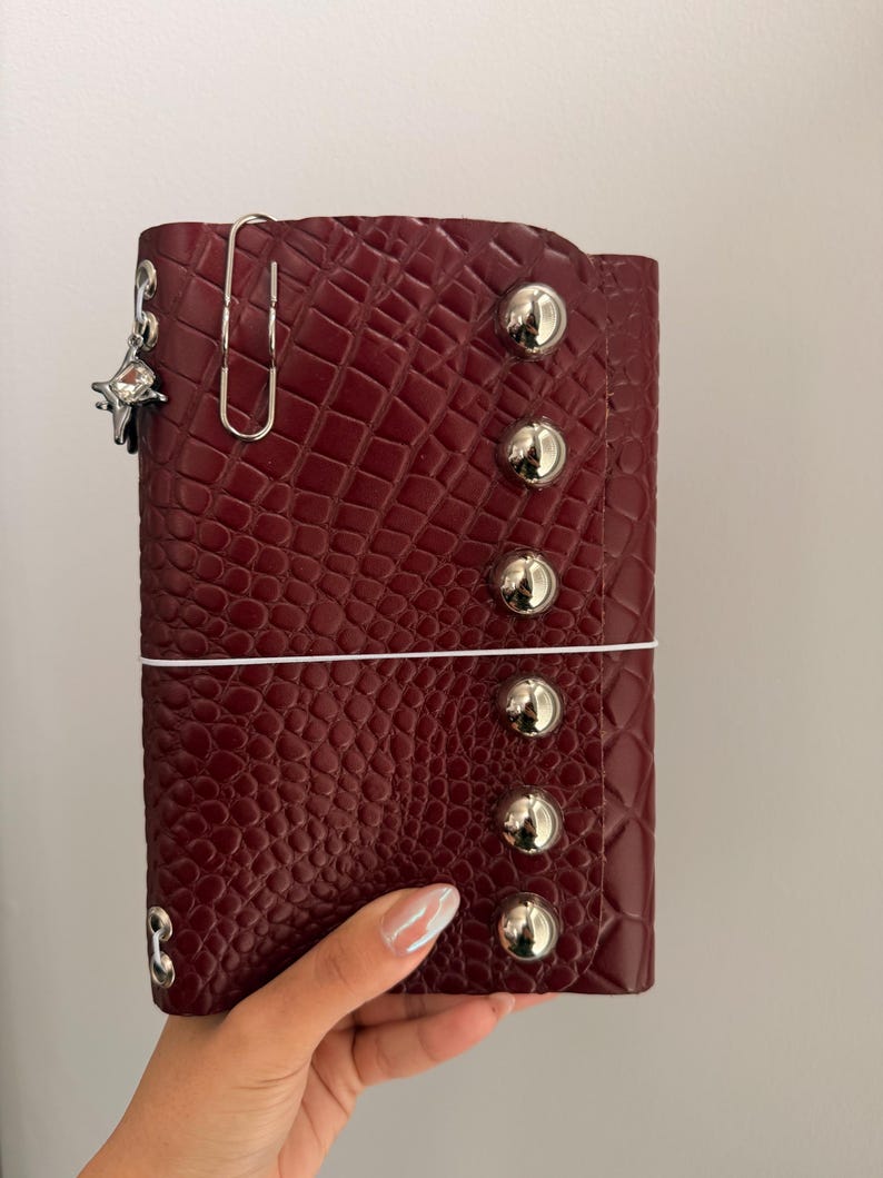 May include: A burgundy faux-leather planner with a crocodile skin texture. The planner has silver metal button accents and a silver paper clip. A small star-shaped charm hangs from the planner.