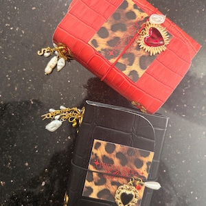May include: Two leather journals, one red and one black, with a crocodile texture. Each journal features a leopard print accent and a heart-shaped charm. The red journal has a red heart charm, while the black journal has a gold heart charm.
