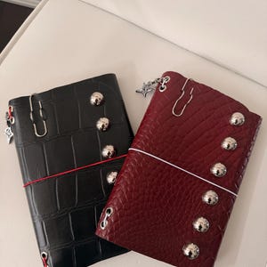 May include: Two leather journals, one black and one burgundy, with a crocodile texture. Each journal features silver-colored studs, a decorative charm, and a paper clip closure. A red elastic band secures the black journal.