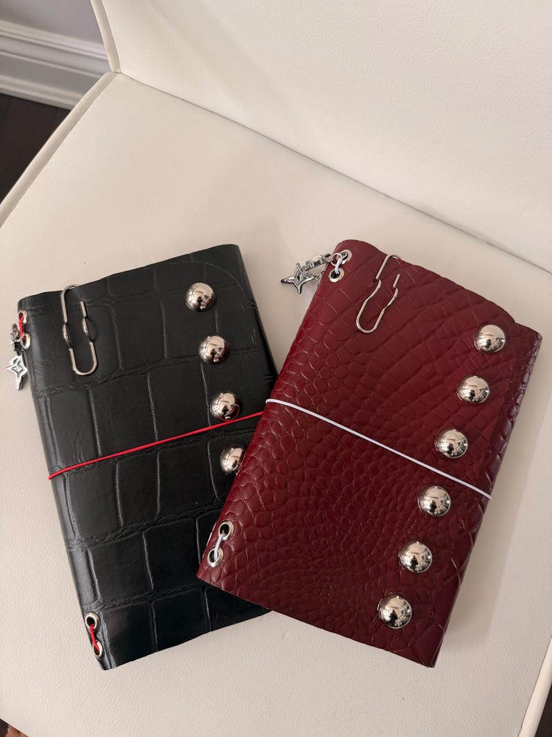 May include: Two leather journals, one black and one burgundy, with a crocodile texture. Each journal features silver-colored studs, a decorative charm, and a paper clip closure. A red elastic band secures the black journal.