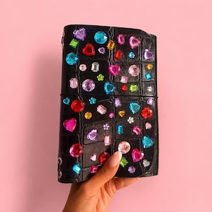 May include: A black notebook with a textured crocodile skin pattern, decorated with numerous colourful plastic rhinestones in various shapes including hearts, circles, and squares in shades of pink, blue, red, yellow, and purple.