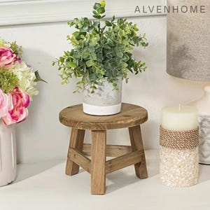 May include: A small, round, wooden plant stand with three legs supports a potted plant with green foliage. The plant pot is white and gray. A white candle with a rope and stone base sits nearby. The word "ALVENHOME" is visible.