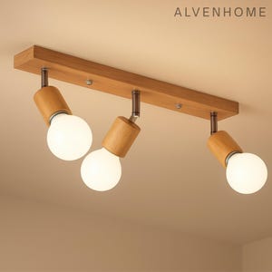 May include: A wooden track light fixture with three adjustable spotlights. The fixture has a rectangular wooden base with three cylindrical wooden light heads. Each head holds a round, white light bulb. The fixture is mounted on a light-colored ceiling.