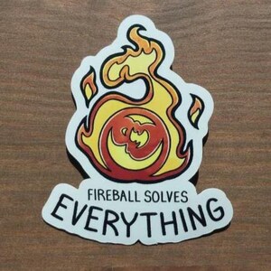 May include: A sticker featuring a flame design in yellow, orange, and red, with the text "Fireball Solves Everything" in black. The sticker has a white border and is on a wooden surface.