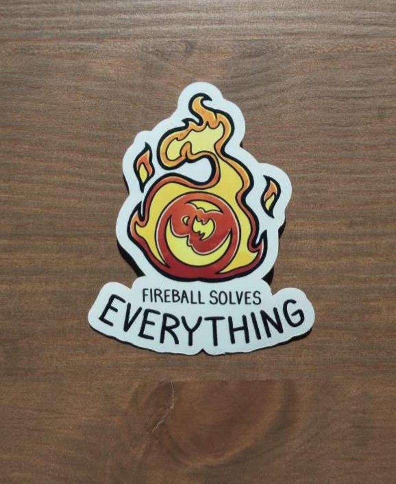 May include: A sticker featuring a flame design in yellow, orange, and red, with the text "Fireball Solves Everything" in black. The sticker has a white border and is on a wooden surface.