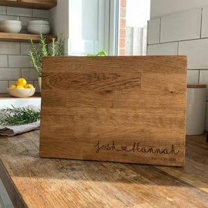 May include: A rectangular wooden chopping board with a light brown finish. The board is engraved with the names "Josh & Hannah" and the date "Est. 11.21.2020". It rests on a wooden countertop in a kitchen setting.