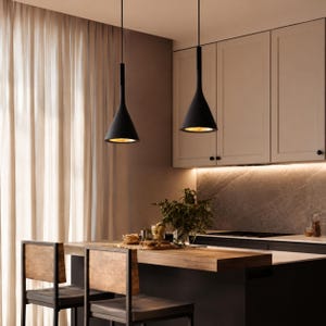 Könnte beinhalten: Zwei schwarze Pendelleuchten mit konischem Schirm hängen über einer Frühstücksbar aus Holz. Die Leuchten haben eine goldene Innenseite und sind von der Decke abgehängt. Die Küche verfügt über weiße Schränke, eine Marmorrückwand und Holzstühle.