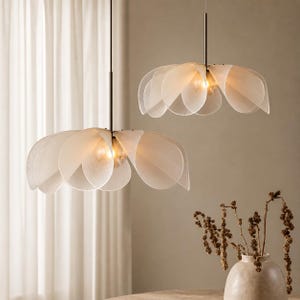Peut inclure: Deux suspensions lumineuses en forme de fleur. Chaque luminaire est composé de pétales translucides entourant une ampoule centrale. Elles sont suspendues à des tiges noires, diffusant une lumière douce. En arrière-plan, un rideau transparent et un vase.