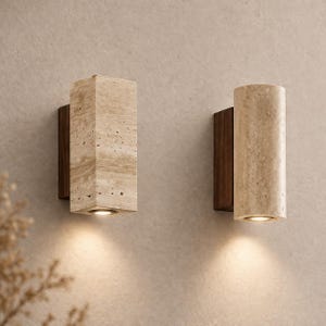 Può includere: Due applique da parete, una rettangolare e una cilindrica, realizzate in pietra beige e montate su legno scuro. Ogni applique ha un'unica fonte di luce nella parte inferiore, che emette un bagliore caldo. Lo sfondo è beige neutro.