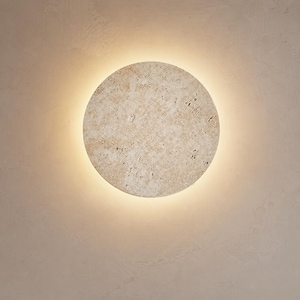 Può includere: Un'applique da parete rotonda beige con una superficie strutturata. La lampada emette una luce calda e soffusa che crea un effetto alone attorno al perimetro. L'apparecchio è montato su una parete dai toni neutri.