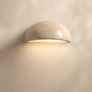 Può includere: Applique da parete a forma di semicerchio, di colore chiaro, con superficie strutturata. L'applique emette una luce calda e invitante dalla sua base curva. L'apparecchio è montato su una parete dai toni neutri.