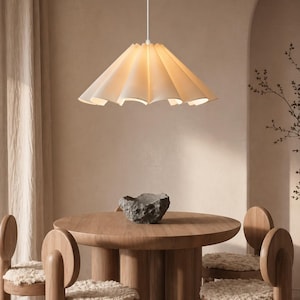 Peut inclure: Un abat-jour en papier plissé de couleur crème est suspendu au-dessus d'une table à manger en bois. La table est entourée de chaises en bois avec des coussins de siège crème. Un bol décoratif en pierre est posé sur la table.