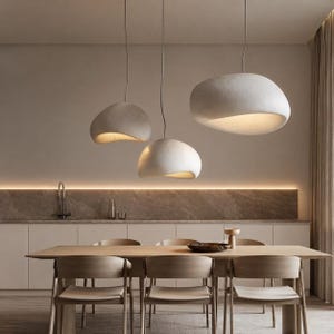 Puede incluir: Tres lámparas colgantes modernas, de color blanco roto, con formas orgánicas, cuelgan sobre una mesa de comedor de madera. Las luces tienen una superficie texturizada y emiten un brillo cálido. El juego de comedor incluye sillas de madera.