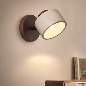 Può includere: Un moderno faretto da parete. Presenta una testa della lampada cilindrica bianca e una base circolare marrone scuro. La lampada è angolata verso il basso, illuminando la parete. Progettato per uso interno.