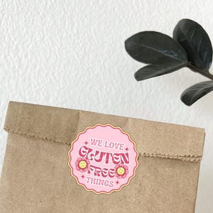 May include: A brown paper bag with a pink and white sticker that reads "WE LOVE GLUTEN FREE THINGS." The sticker has a scalloped edge and flower accents. The bag is sealed at the top. A plant with dark green leaves is in the background.