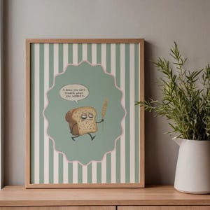 Puede incluir: Una impresión artística enmarcada con una ilustración de dibujos animados de una rebanada de pan tostado con una expresión triste, sosteniendo una espiga de trigo. El texto en un bocadillo dice: "Sabía que eras un problema cuando entraste".