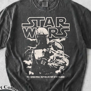 Puede incluir: Camiseta gris oscuro con un gr&aacute;fico de The Mandalorian y Grogu de Star Wars. El logotipo de Star Wars est&aacute; encima de la imagen, con texto debajo. La camiseta es una camiseta Comfort Colors Pepper.