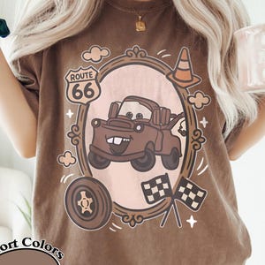 May include: Espresso-colored t-shirt with a cartoon graphic of a brown tow truck inside a decorative frame. The design includes Route 66, checkered flags, and a tire. The shirt is labeled "Comfort Colors".