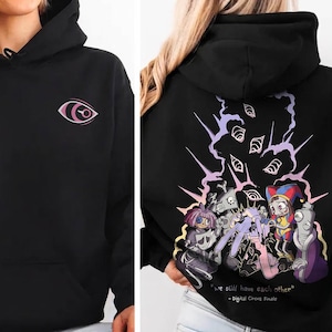 May include: A black hooded sweatshirt featuring a small pink eye graphic on the front chest. The back displays a colorful illustration of animated characters with multiple eyes above them and the text "we still have each other" and "Digital Circus Finale".