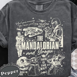 May include: Dark grey Comfort Colors t-shirt with a graphic design featuring characters and text from The Mandalorian series. The design includes the text "Mind your manners, You know what I'm talking about." and "The Mandalorian and Grogu."