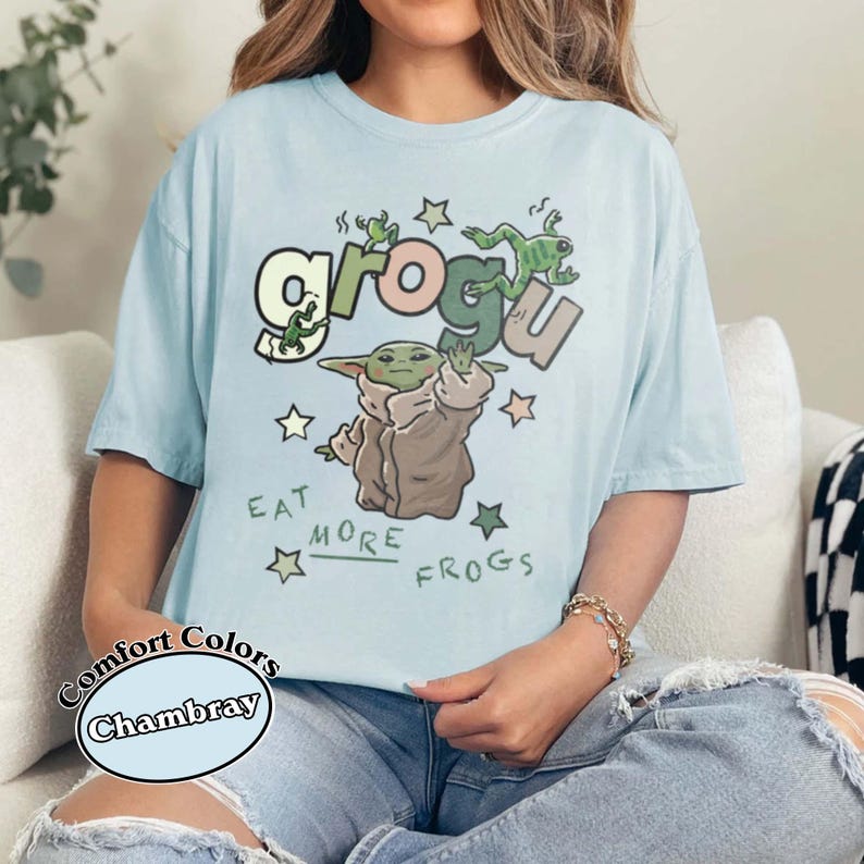 May include: Light blue Comfort Colors chambray t-shirt featuring a Grogu graphic with the text "Eat More Frogs." The design includes Grogu, frogs, and stars. The shirt is a relaxed fit.