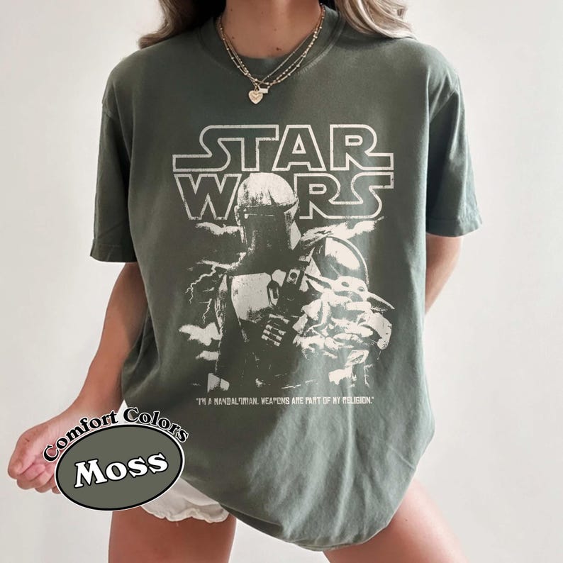 Puede incluir: Camiseta verde musgo con un dise&ntilde;o de Star Wars, que incluye el texto "STAR WARS" y una imagen de The Mandalorian y Grogu. La camiseta tambi&eacute;n incluye la cita, "I'M A MANDALORIAN, WEAPONS ARE PART OF MY RELIGION." La camiseta es de Comfort Colors.