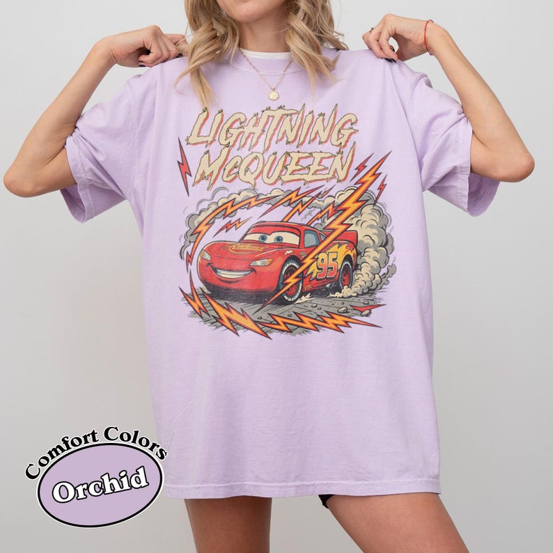 May include: Orchid-colored t-shirt featuring a graphic of Lightning McQueen from the Cars movie. The design includes the text "Lightning McQueen" and the number "95" with lightning bolt accents. The shirt is a Comfort Colors brand.