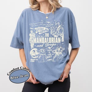 May include: Blue jean coloured t-shirt with a graphic featuring characters and text from "The Mandalorian" series. The design includes the text "The Mandalorian and Grogu", along with other phrases and images from the show.