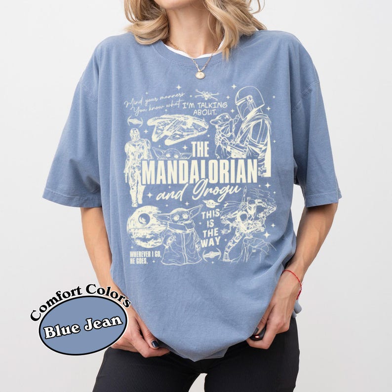 May include: Blue jean coloured t-shirt with a graphic featuring characters and text from "The Mandalorian" series. The design includes the text "The Mandalorian and Grogu", along with other phrases and images from the show.