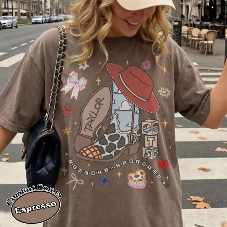 Puede incluir: Una camiseta color espresso con un estampado gr&aacute;fico de una bota vaquera con patr&oacute;n de vaca, un sombrero rojo, un lazo rosa y bloques apilados con las letras T, S y 13. El dise&ntilde;o incluye peque&ntilde;as flores, estrellas y un collar de cuentas.