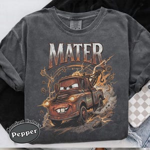 May include: Dark gray sweatshirt featuring a graphic of Mater from the movie Cars. The design includes the word "MATER" above the character, with lightning bolts and tools. The sweatshirt has the words "Comfort Colors Pepper" printed on the bottom left.