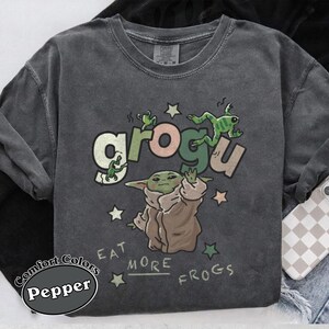 May include: Dark grey Comfort Colors sweatshirt with a Grogu graphic. The design features Grogu with the text "grogu" and "Eat More Frogs", along with green frogs and stars. The sweatshirt is a Pepper color.