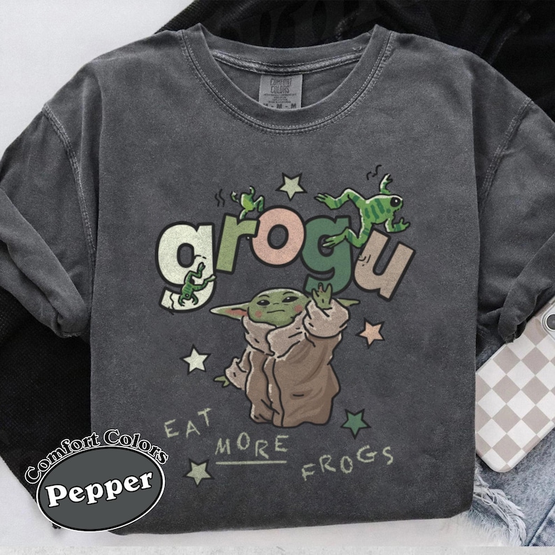 May include: Dark grey Comfort Colors sweatshirt with a Grogu graphic. The design features Grogu with the text "grogu" and "Eat More Frogs", along with green frogs and stars. The sweatshirt is a Pepper color.