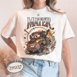 May include: Ivory-colored t-shirt featuring a cartoon image of Mater, the tow truck character. The word "MATER" is printed above the image in a distressed style. The shirt has a Comfort Colors label.