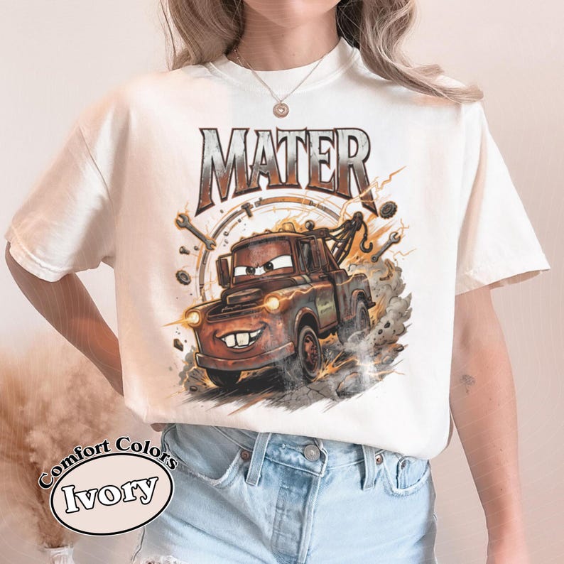 May include: Ivory-colored t-shirt featuring a cartoon image of Mater, the tow truck character. The word "MATER" is printed above the image in a distressed style. The shirt has a Comfort Colors label.