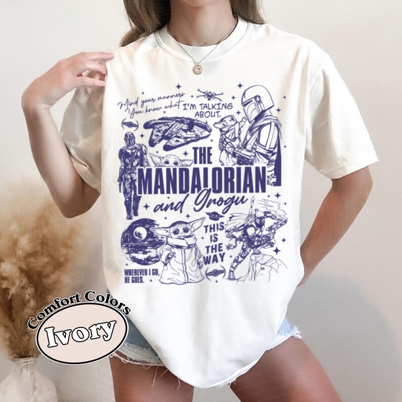 May include: Ivory t-shirt with a blue graphic design featuring characters and phrases from The Mandalorian series. The design includes the words "The Mandalorian and Grogu" and other phrases, along with character illustrations.