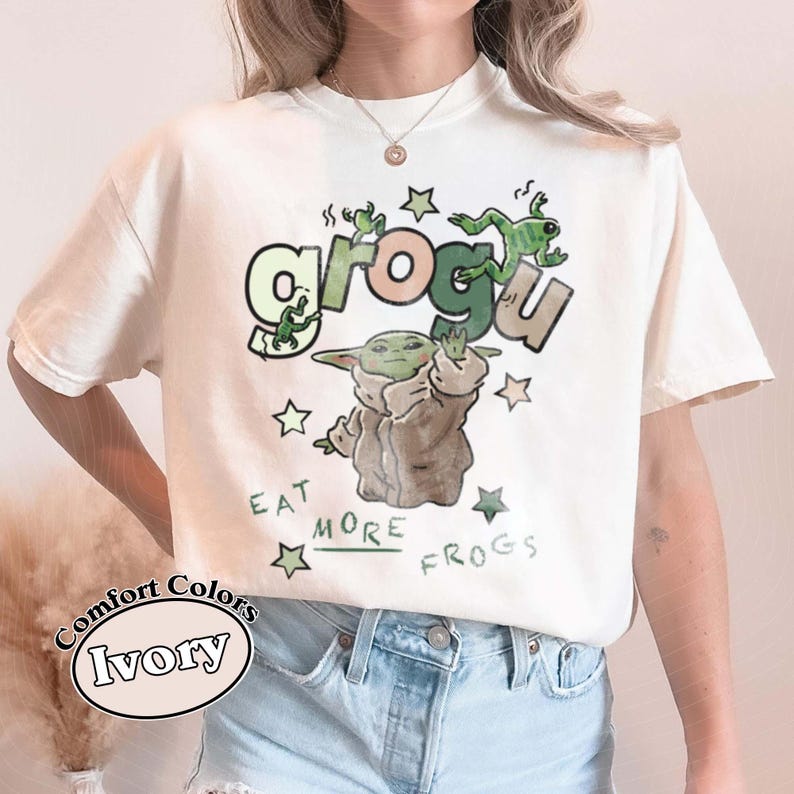 May include: Ivory Comfort Colors t-shirt featuring a Grogu graphic with the text "Eat More Frogs." The design includes Grogu, green frogs, and stars. The shirt is a relaxed fit.