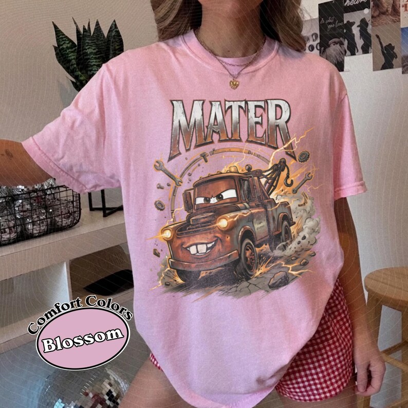 May include: Pink Comfort Colors t-shirt featuring a graphic of Mater from the movie Cars. The design includes the word "MATER" in a vintage font above the character. The shirt has short sleeves and a round neckline.