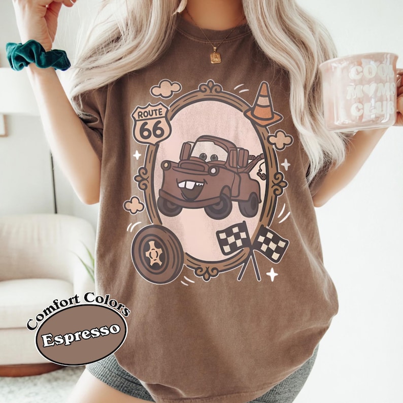 May include: Espresso-colored t-shirt with a cartoon graphic of a brown tow truck inside a decorative frame. The design includes Route 66, checkered flags, and a tire. The shirt is labeled "Comfort Colors".