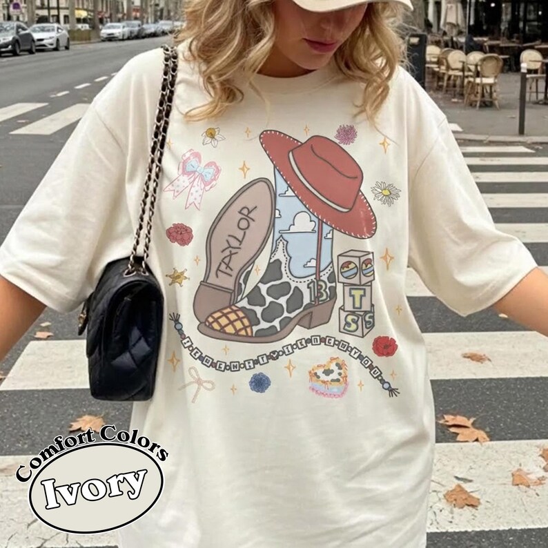 Puede incluir: Una camiseta color marfil con un estampado gr&aacute;fico de una bota vaquera con manchas de vaca, un sombrero rojo, un lazo, flores y bloques con las letras T, S y el n&uacute;mero 13. El dise&ntilde;o incluye una cadena de cuentas con texto y el nombre Taylor.