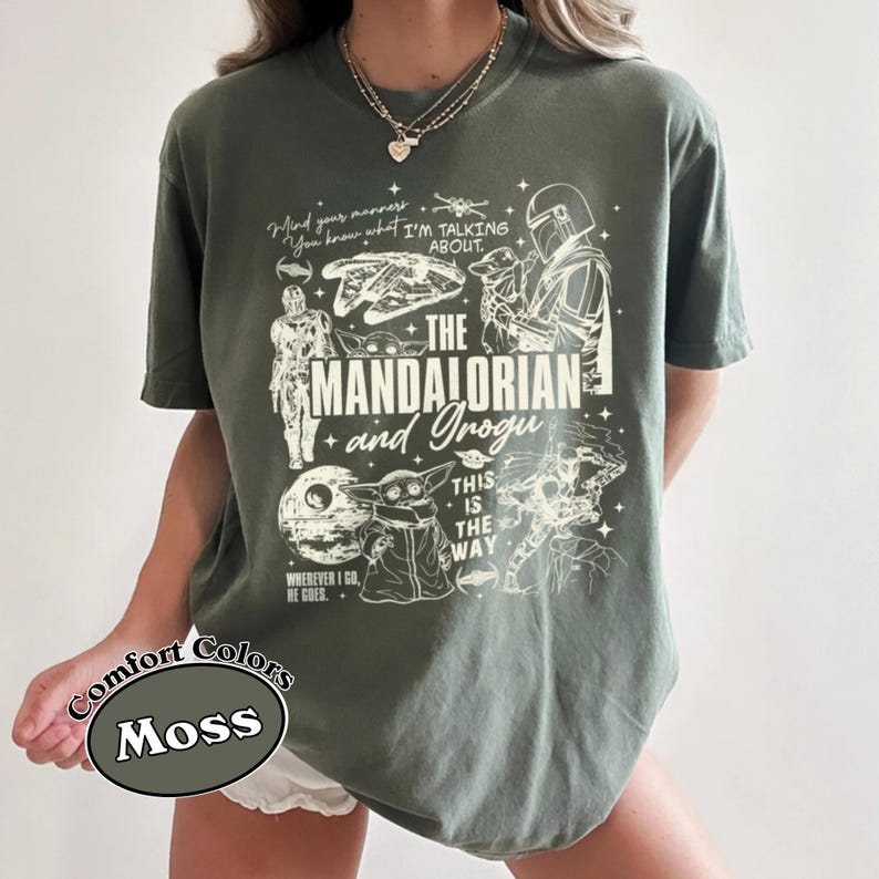 May include: A moss green t-shirt with a graphic design from "The Mandalorian" series. The design includes the words "The Mandalorian and Grogu", along with other phrases and character images. The shirt is labeled "Comfort Colors".