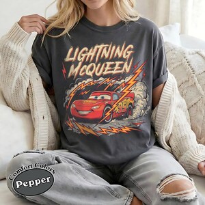 May include: Dark gray t-shirt featuring Lightning McQueen from the Cars movie. The design includes the text "Lightning McQueen" in a distressed font, with the character in red and yellow lightning bolts. The shirt is a Comfort Colors Pepper tee.