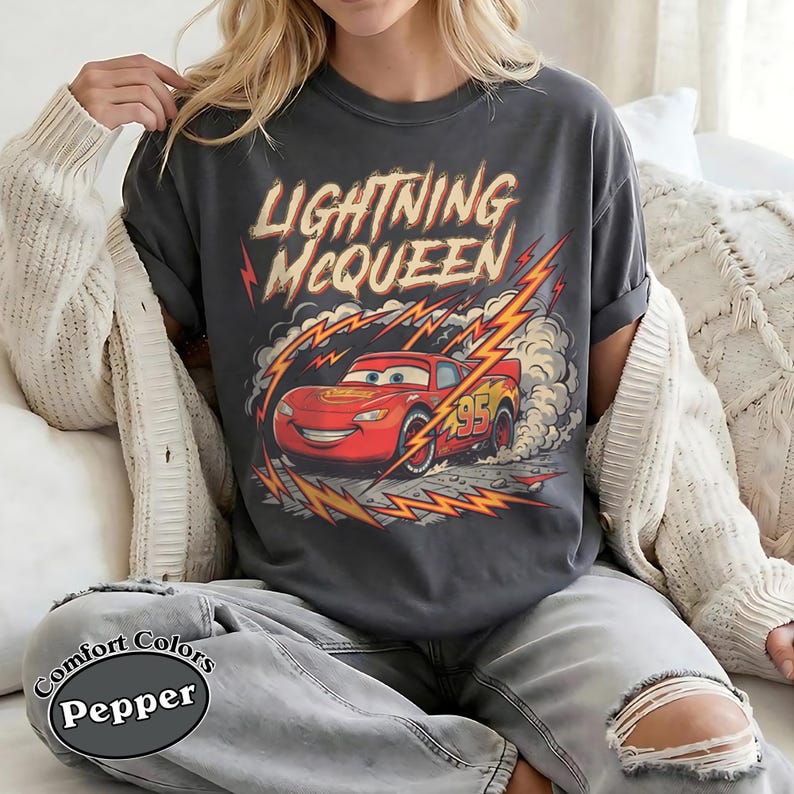 May include: Dark gray t-shirt featuring Lightning McQueen from the Cars movie. The design includes the text "Lightning McQueen" in a distressed font, with the character in red and yellow lightning bolts. The shirt is a Comfort Colors Pepper tee.